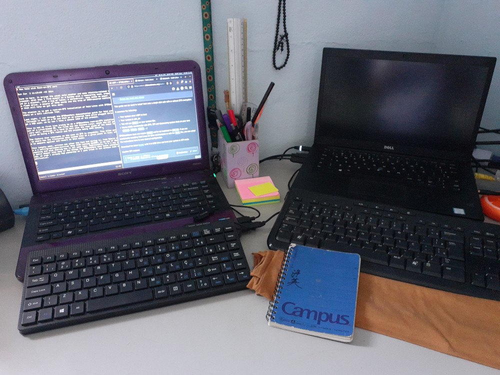 Two laptops side-by-side on a desk, each with a USB keyboard plugged in. Writing utensils inside a holder and post-its are between the two. A sunflower-patterned keychain and prayer beads hang from the wall. The computer on the left has a brown cloth for a wrist rest in front of its USB keyboard, to the left of which lies a small blue Campus notebook.